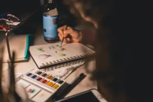 A person painting small colorful objects in a sketchbook using watercolors, surrounded by art supplies on a desk.
